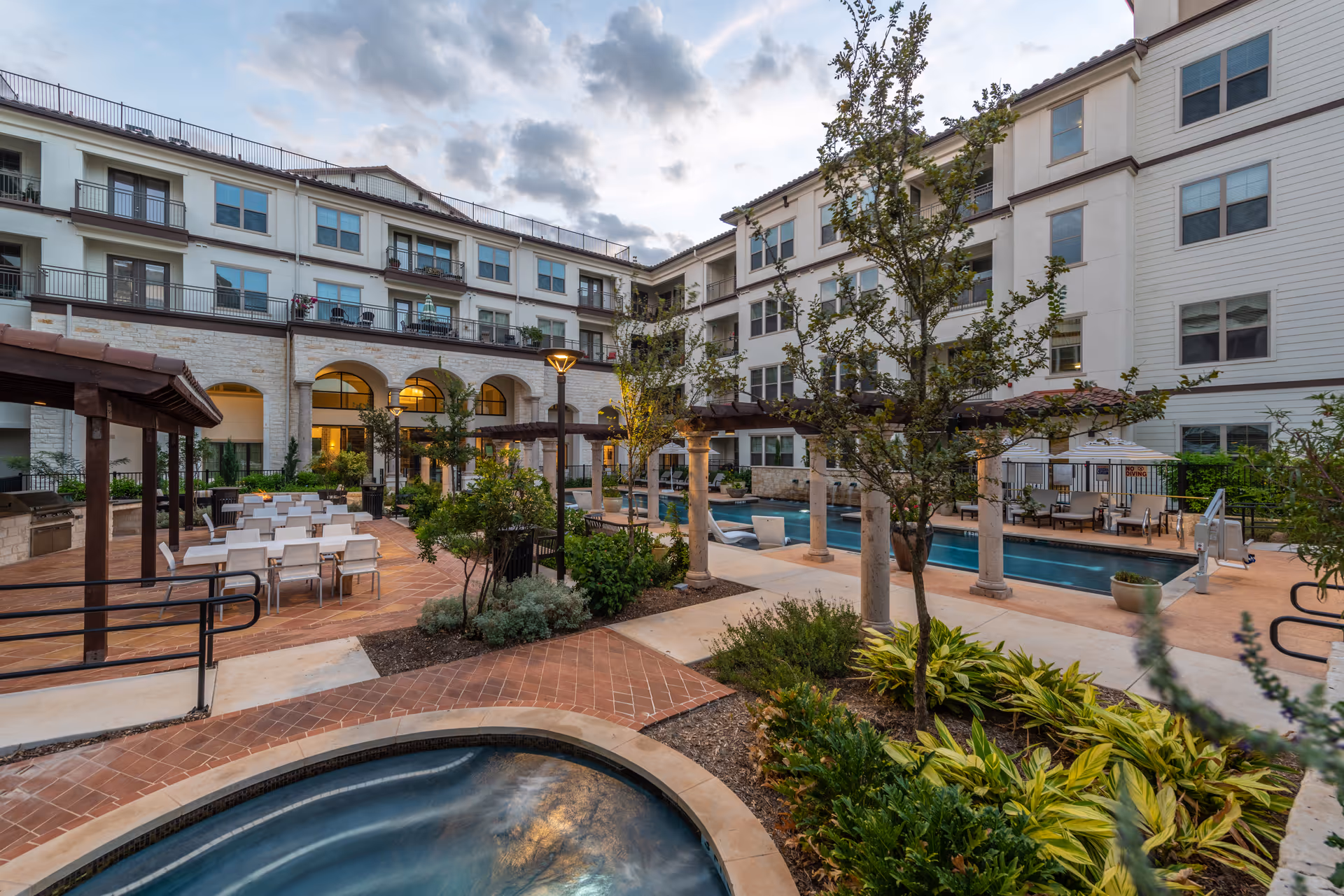 Outdoor courtyard area of Overture Stone Oak featuring a hot tub in the foreground, a swimming pool with lounge chairs, pergolas, dining tables with chairs, landscaped plants, and a multi-story residential building surrounding the courtyard under a partly cloudy sky.