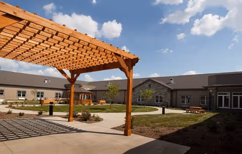 Sunny courtyard with a wooden pergola, pathways, landscaping and the facility building under a blue sky.