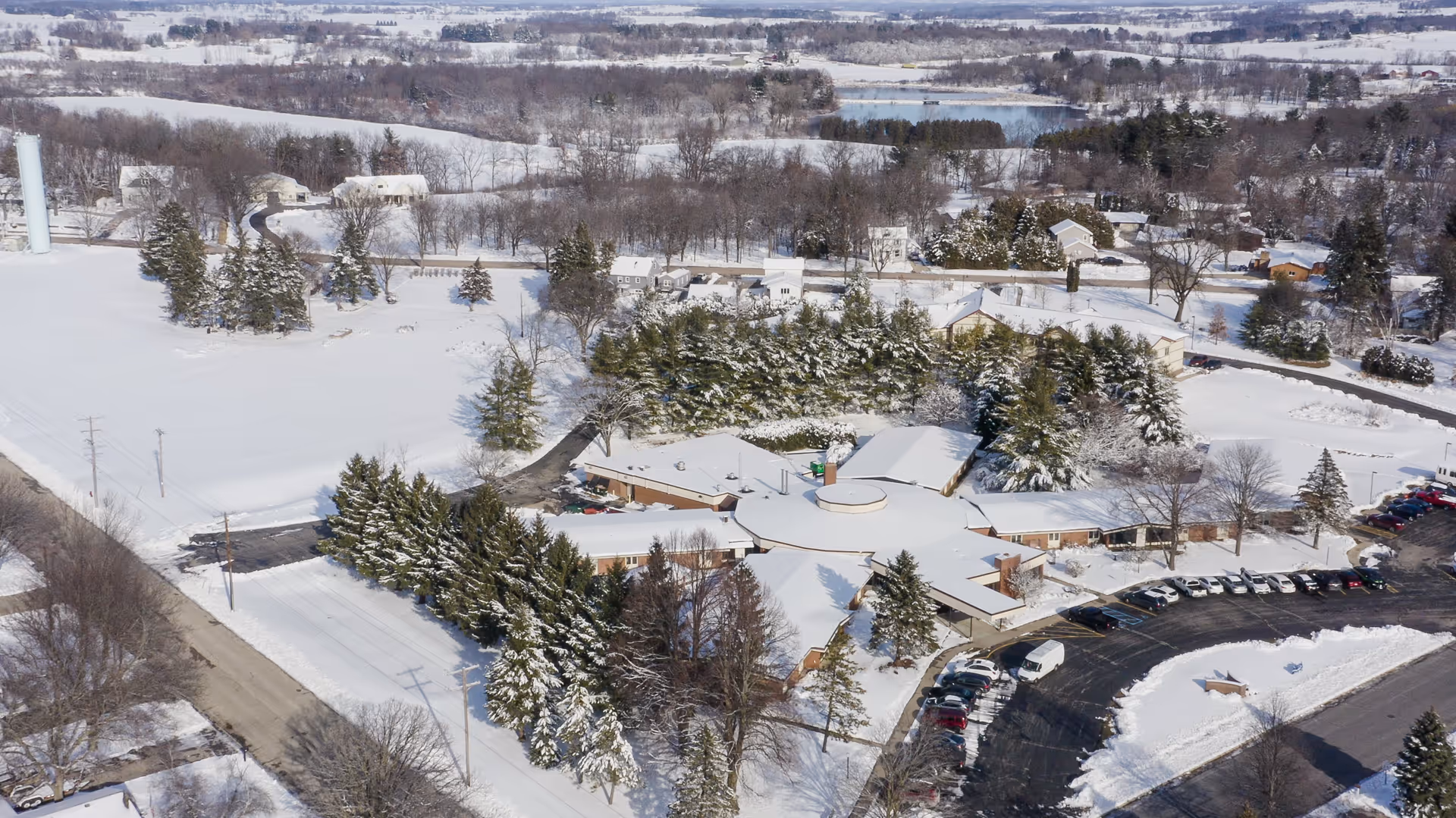 Aerial view of Evansville Manor Skilled Nursing facility surrounded by snow-covered grounds, trees, and nearby roads. The building has a unique star-shaped layout with a circular central section and multiple wings extending outward. Several cars are parked in the parking lot adjacent to the building, and the surrounding landscape is covered in snow with scattered trees and houses in the distance.