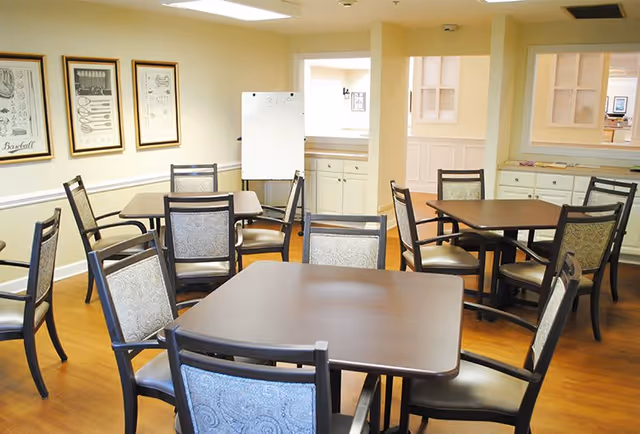 A bright dining room with several square wooden tables and cushioned chairs arranged neatly. The room has light-colored walls adorned with framed artwork and a whiteboard in the background. There is a window-like opening in the wall showing a glimpse of another room with cabinets.