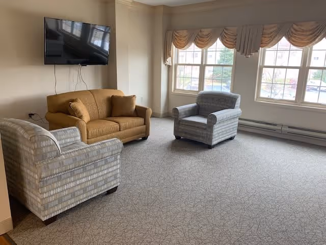 A living room area with a brown loveseat and two patterned armchairs arranged on a carpeted floor. A flat-screen TV is mounted on the wall above the loveseat. Large windows with beige valances allow natural light to fill the room.