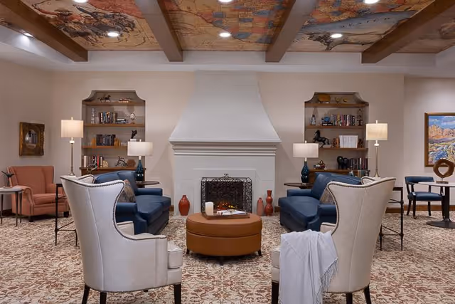 A cozy living room with a central fireplace flanked by built-in bookshelves. The room features two blue sofas facing each other, two beige armchairs with a white throw draped over one, and a round brown ottoman in the center. There are several table lamps providing warm lighting, a patterned carpet, and a decorative ceiling with artwork. Additional seating and framed artwork are visible in the background.