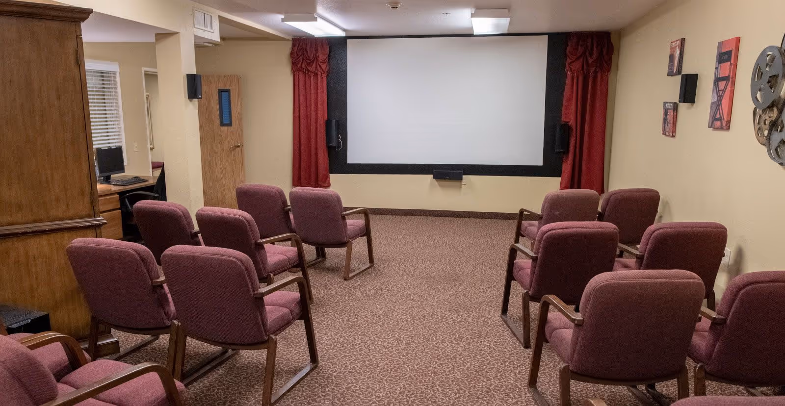 Small screening room with rows of upholstered chairs facing a large projection screen and wall-mounted speakers.