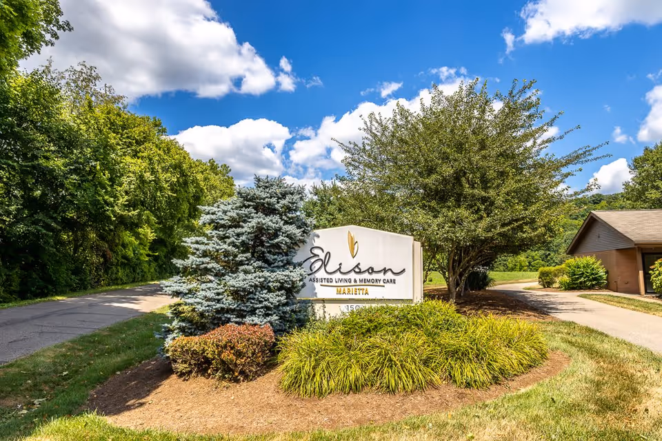 Outdoor view of the entrance sign for Elison Assisted Living & Memory Care of Marietta surrounded by landscaped bushes and trees under a partly cloudy blue sky.
