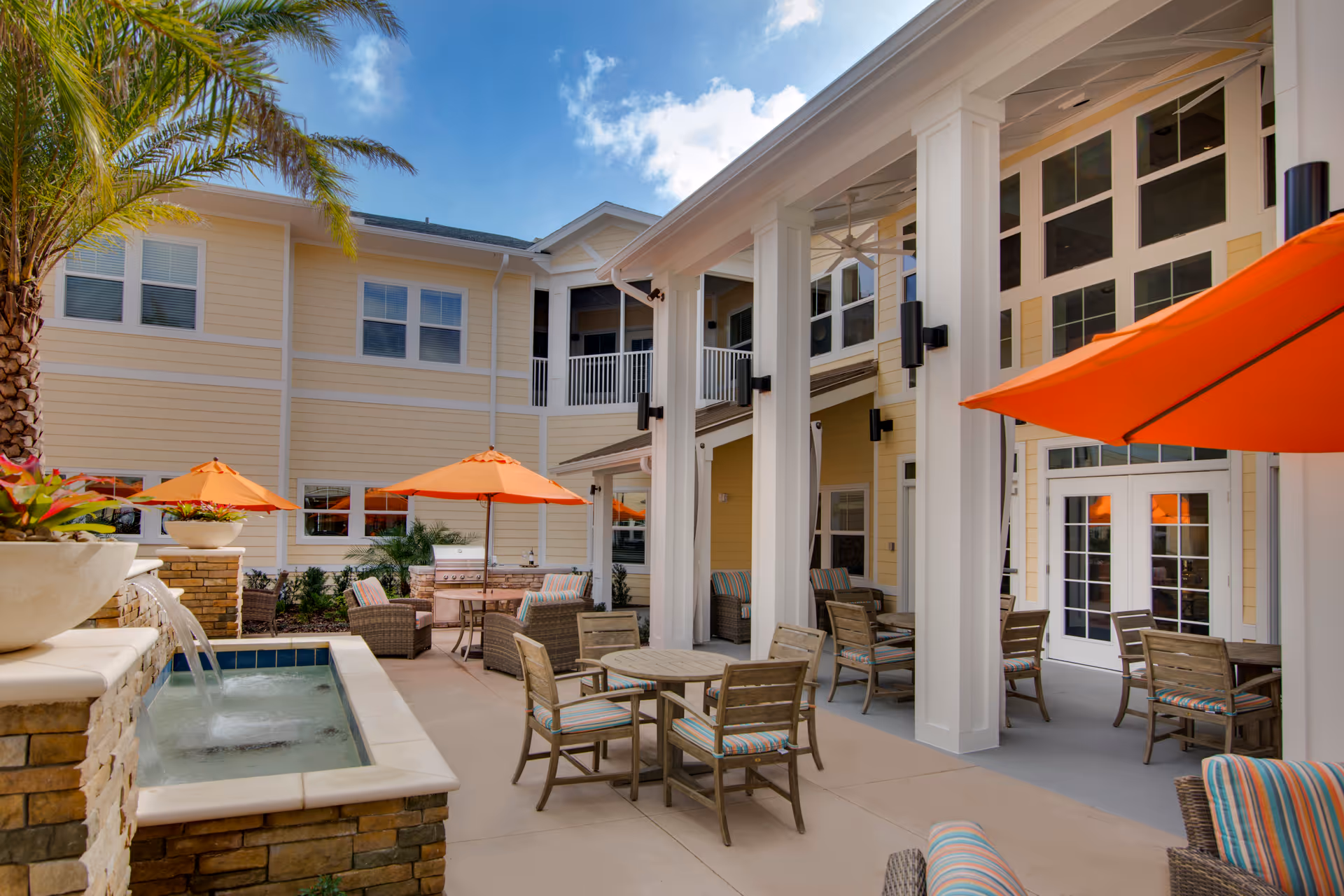 Sunny outdoor courtyard with tables and chairs, orange umbrellas, a small fountain, and a two-story yellow building.