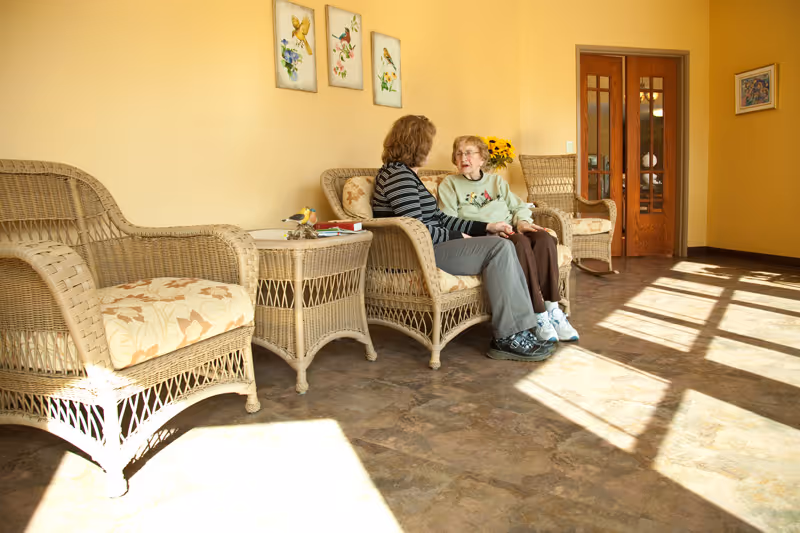 Two women sitting and talking on wicker chairs in a sunlit room with yellow walls, floral artwork on the wall, and a wicker side table between them.
