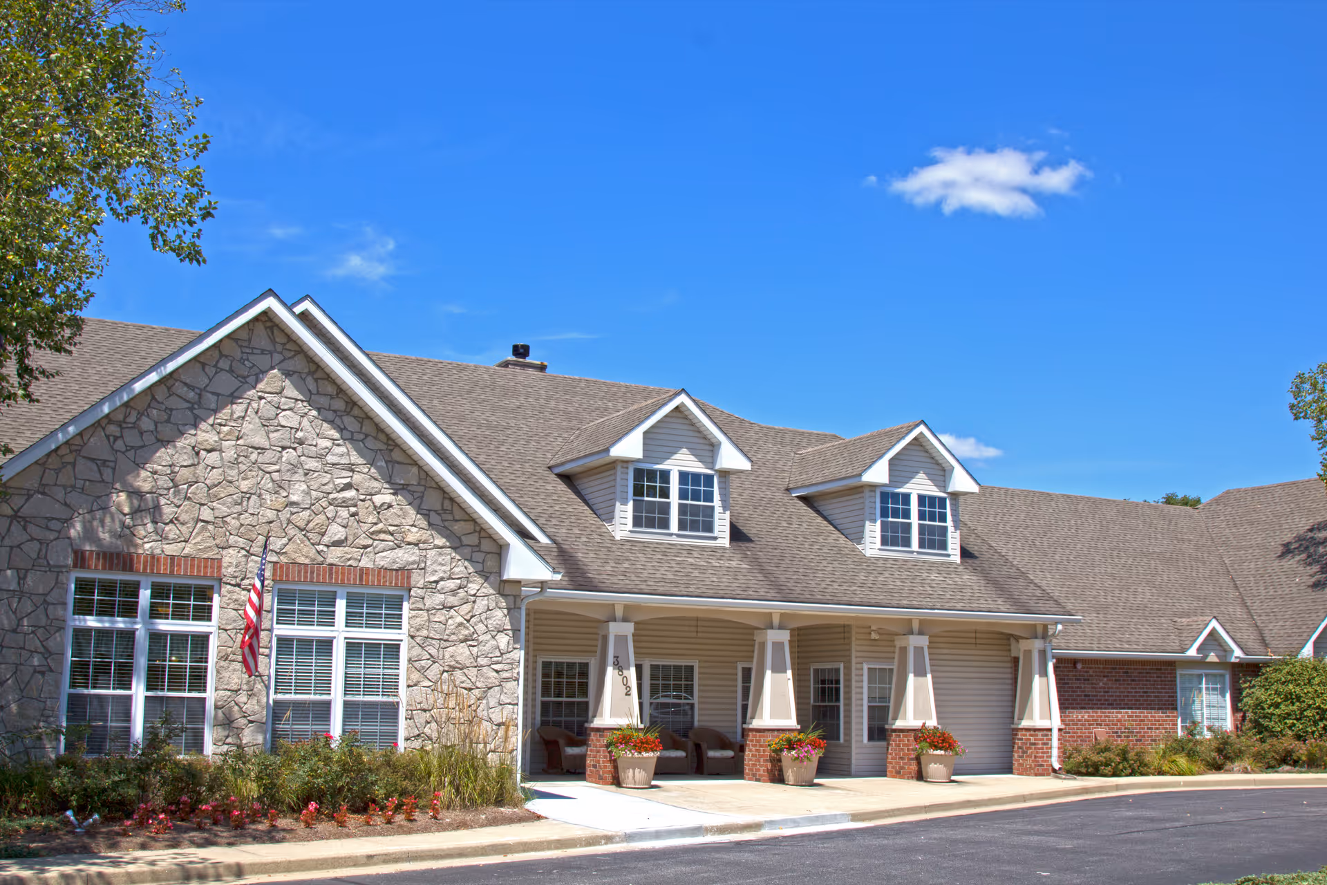 Front exterior of a single-story senior living building with a covered entrance, stone and siding facade, dormer windows, landscaping and an American flag.