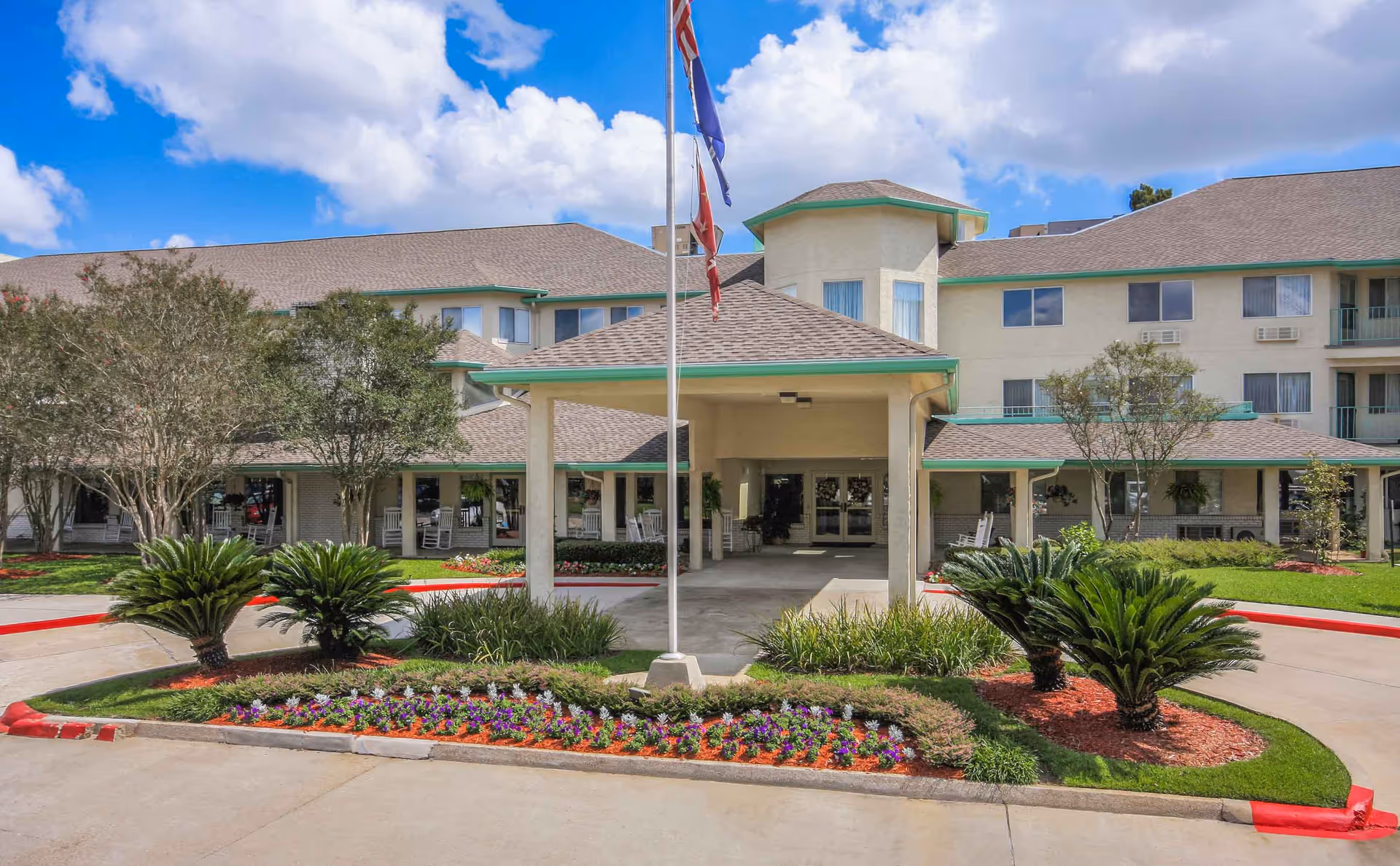 Front exterior view of Whealdon Estates by Barclay House, a multi-story senior living facility with a covered entrance, landscaped flower beds, palm plants, and trees under a partly cloudy blue sky.