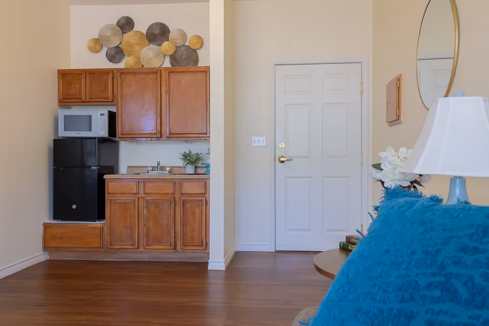 Interior view of a small kitchenette area with wooden cabinets, a black mini refrigerator, and a microwave. There is a sink with a small plant and decorative items on the countertop. To the right, there is a white door, a round mirror on the wall, a table with a white lamp, and a blue cushion on a chair or sofa partially visible.