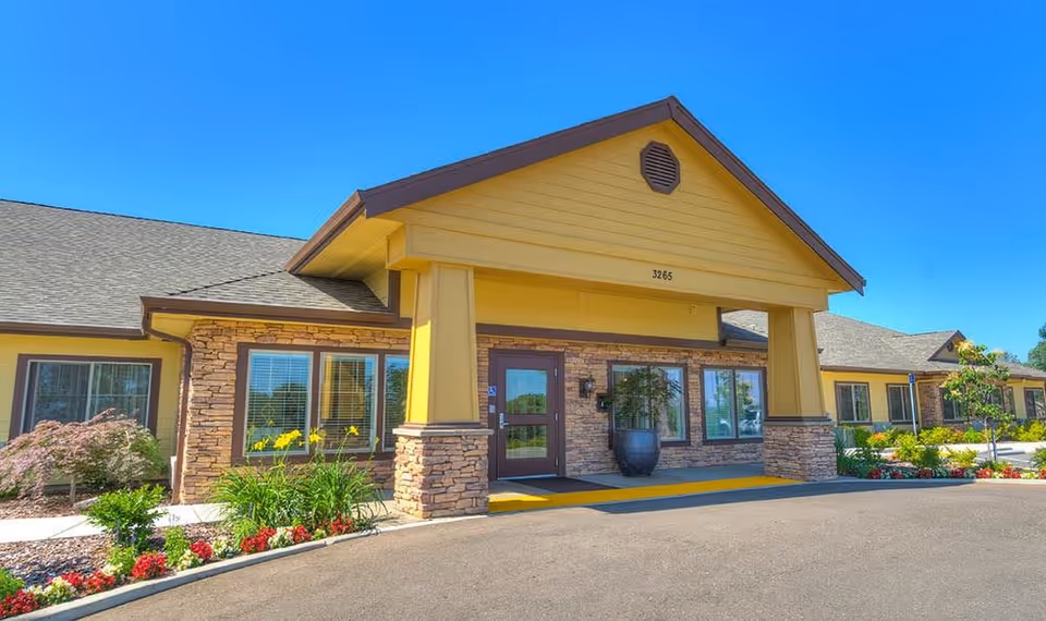 Exterior front view of a single-story building with yellow siding and stone accents, featuring a covered entrance with two large pillars. The building is surrounded by landscaped flower beds and shrubs under a clear blue sky.
