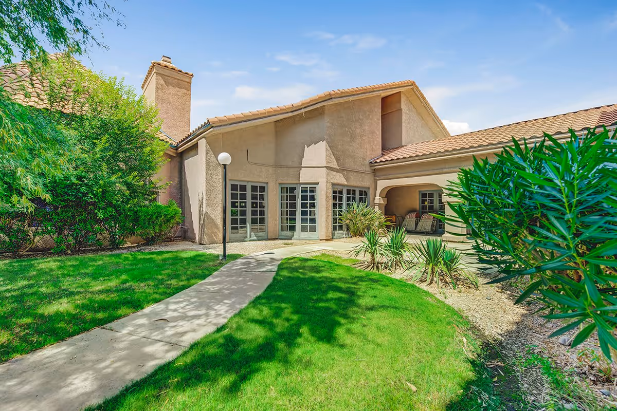 Exterior view of a senior living facility building with beige stucco walls and a tiled roof, surrounded by green grass, bushes, and a concrete pathway leading to the entrance. The sky is clear and blue.