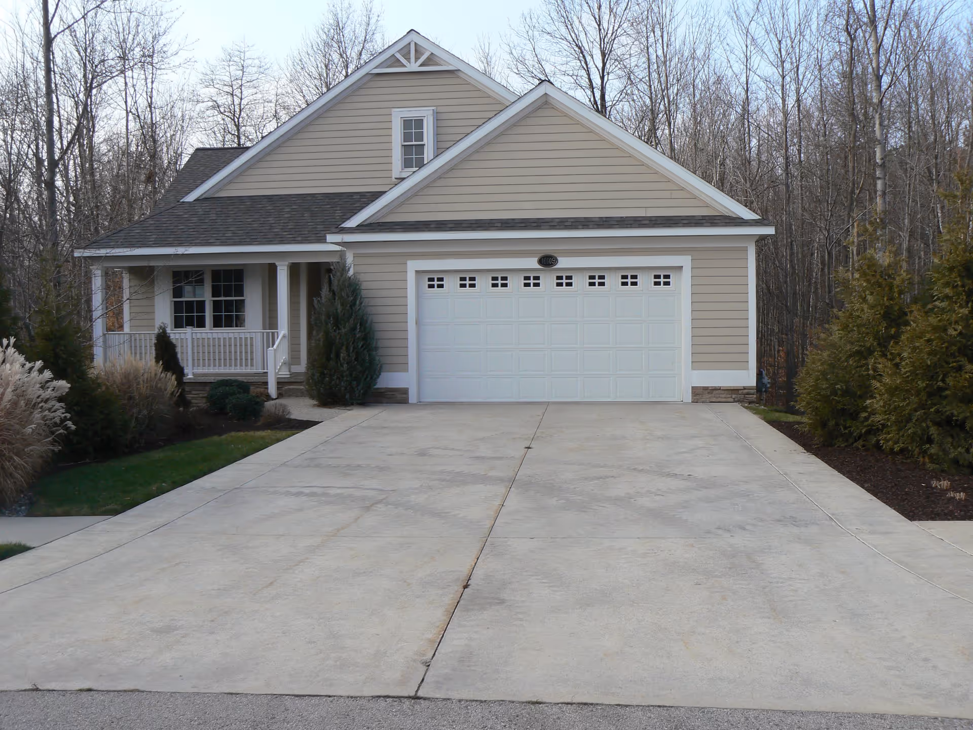 Front exterior view of a beige single-family house with a two-car garage, a small porch with white railings, and a concrete driveway. The house is surrounded by some bushes and trees without leaves in the background.