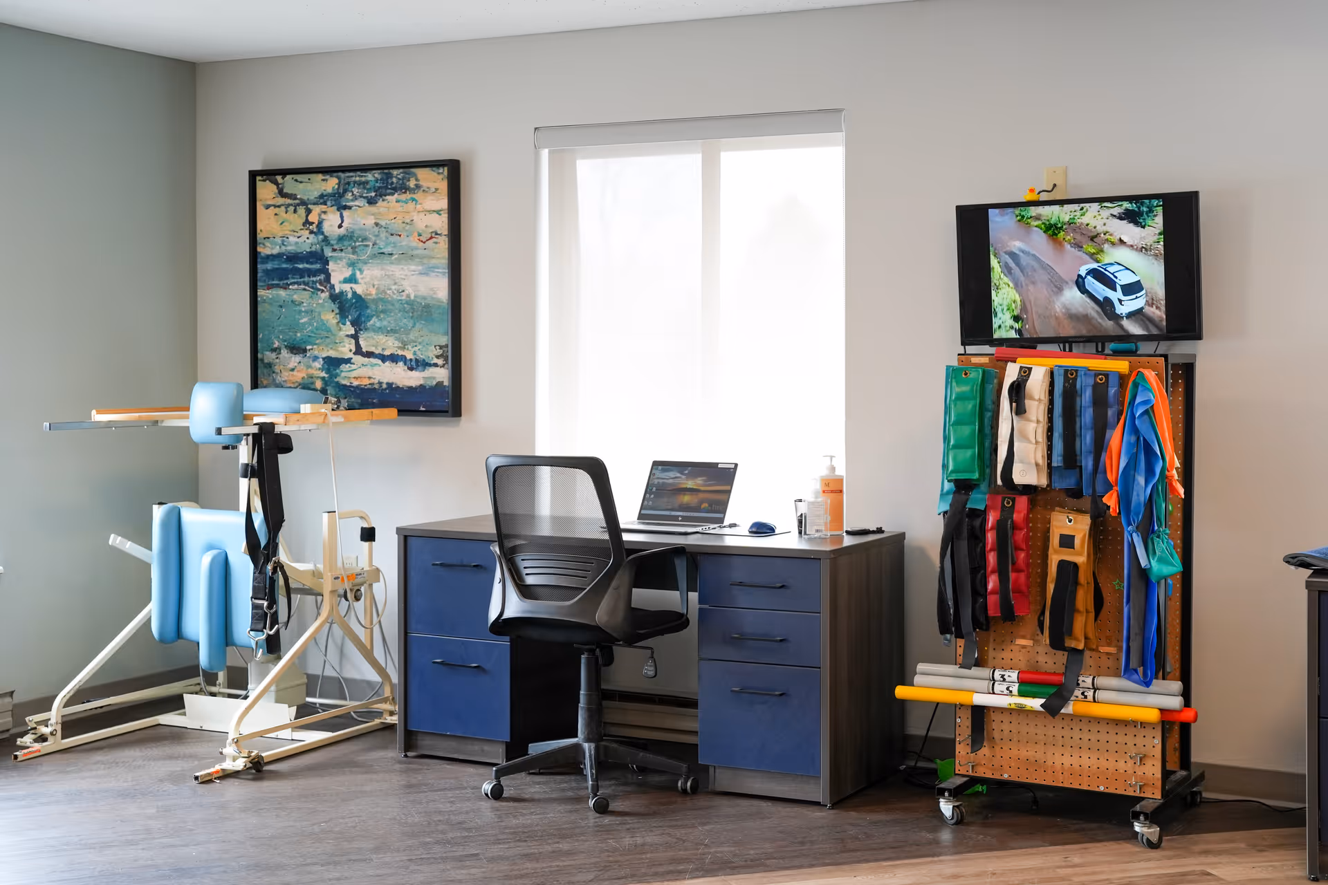 A therapy or rehabilitation room with a blue and white exercise machine on the left, a desk with a black mesh office chair and a laptop in the center, and a pegboard on wheels holding various colored exercise bands and equipment on the right. A TV mounted above the pegboard shows a car driving on a dirt road. A large window with a white blind is behind the desk, and a framed abstract painting hangs on the wall to the left.