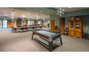 A spacious recreational room with a shuffleboard table in the foreground, a long table with chairs in the background, and a wooden cabinet against the wall. The room has green walls, beige carpet, and ceiling decorations including hanging snowflakes.