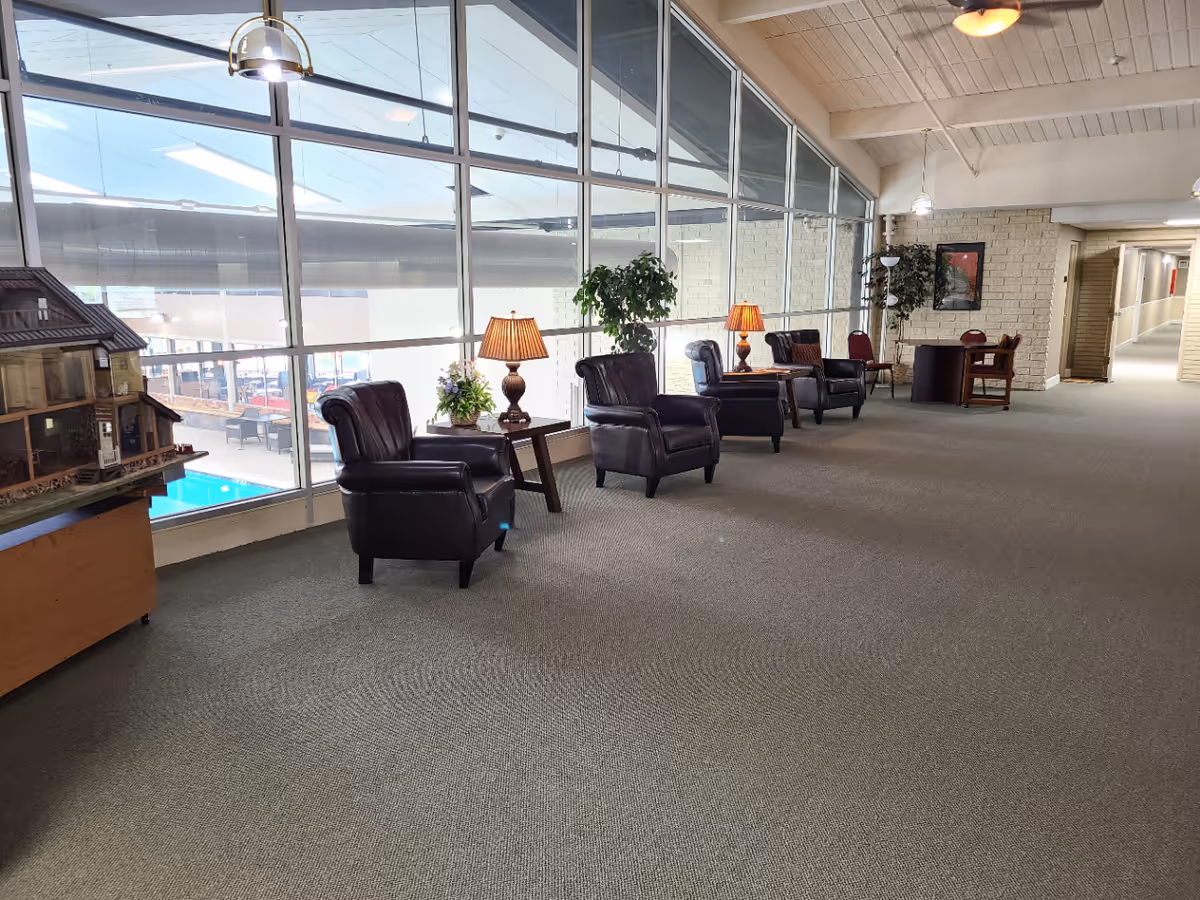Long lounge area with leather armchairs, side tables and lamps along large windows overlooking an indoor pool.
