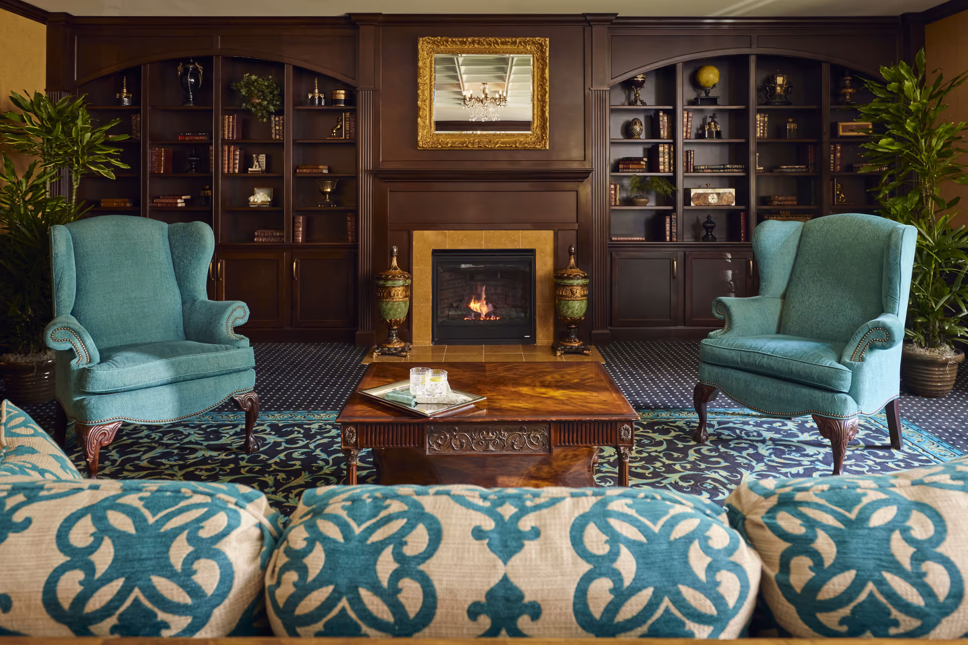 A cozy living room with two teal upholstered armchairs and a patterned sofa facing a wooden coffee table. Behind the table is a fireplace with a fire burning, flanked by two decorative urns. Dark wooden built-in bookshelves filled with books and decorative items line the wall, and a gold-framed mirror hangs above the fireplace. Two large potted plants are placed on either side of the bookshelves.