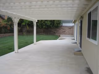 Covered concrete patio beside a beige building, white posts, and a green lawn.