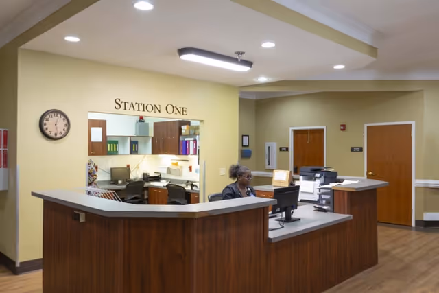 Reception desk area labeled Station One with a woman sitting behind the desk working on a computer. The room has beige walls, wooden doors, and a clock on the wall showing 12:10.