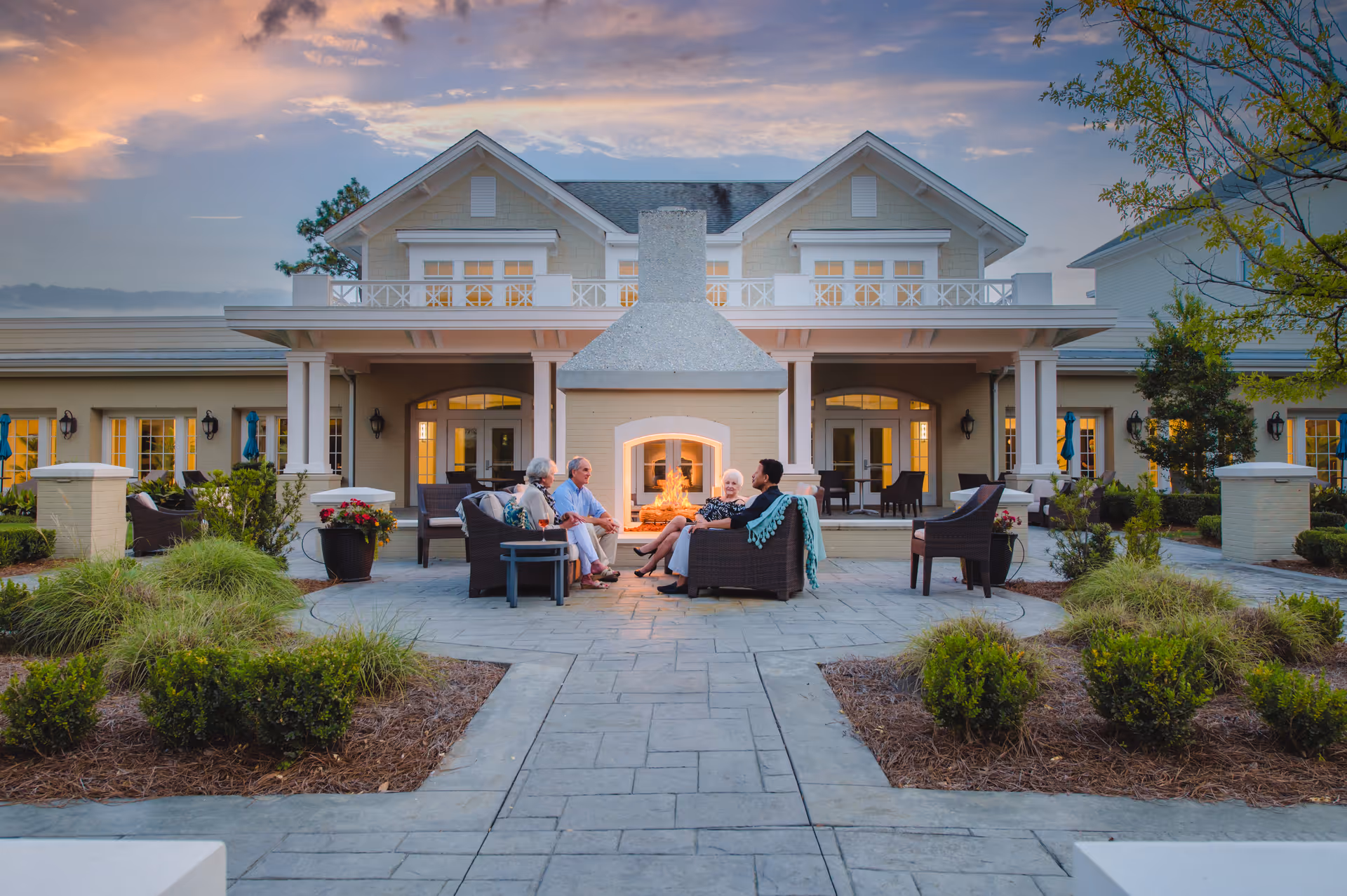 Four elderly people sitting on outdoor wicker chairs around a lit fireplace in front of a large building with a porch and balcony at sunset, surrounded by landscaped bushes and trees.