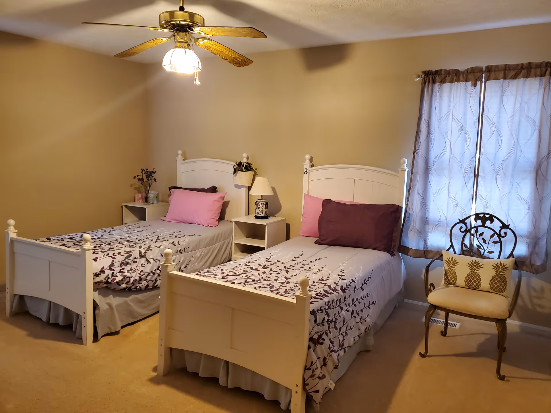 A bedroom with two white twin beds, each with floral-patterned bedding and pink and purple pillows. Between the beds is a small white nightstand with a lamp and a small plant. A decorative chair with a pineapple-patterned cushion is placed near a window with sheer curtains. A ceiling fan with lights is mounted on the ceiling.