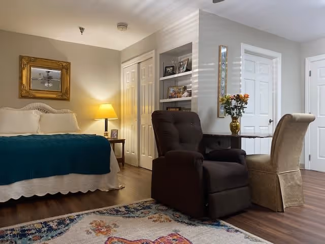 A cozy bedroom with a bed covered in white and teal bedding, a nightstand with a lamp, a large mirror on the wall, a dark cushioned armchair, a beige upholstered chair, a small table with a vase of flowers, built-in shelves with framed photos, and wooden flooring with a colorful area rug.