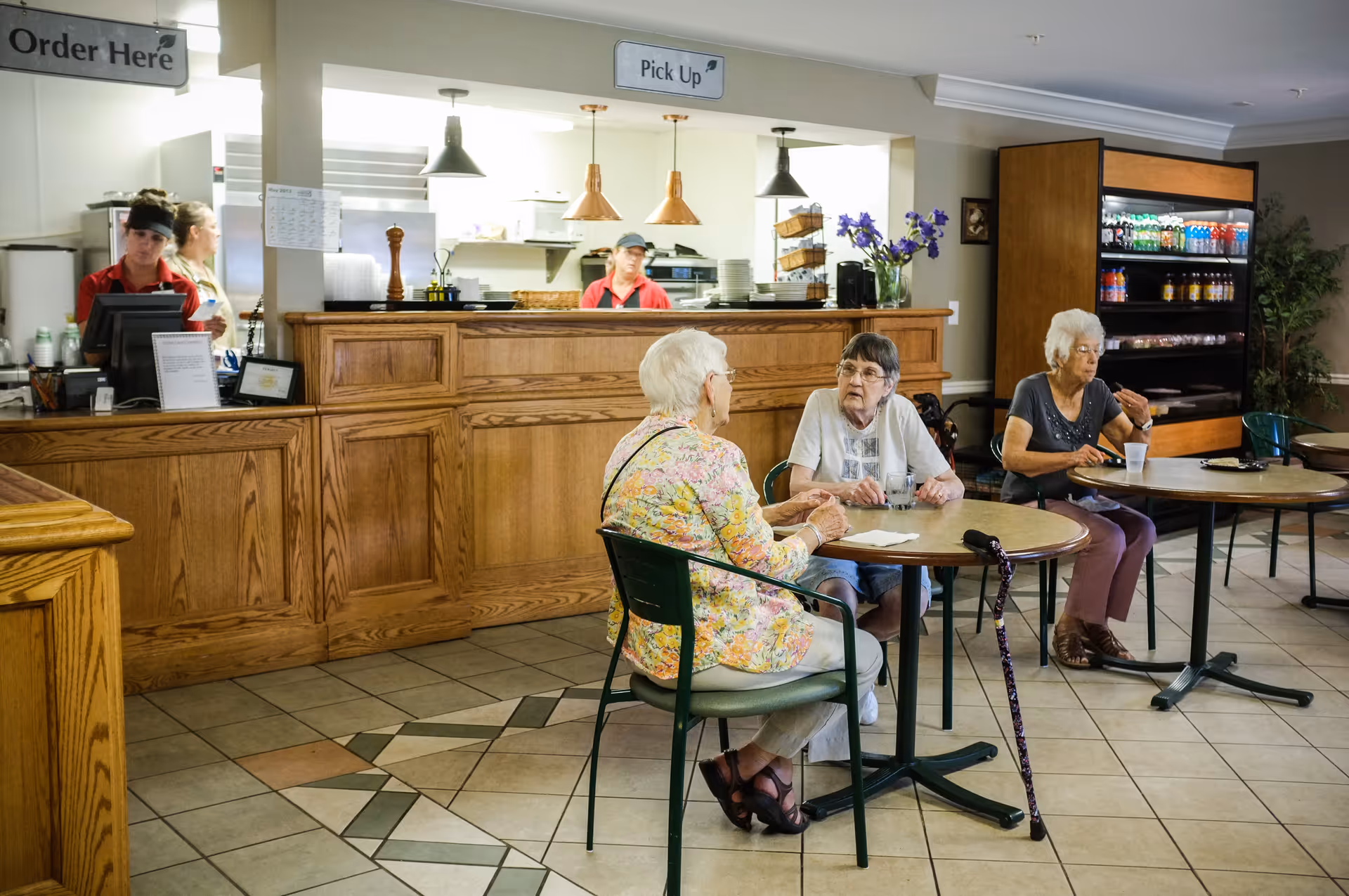 Three elderly women sitting at separate tables in a dining area of a senior living facility. Two women are engaged in conversation at one table, while the third woman is eating at another table. Behind them is a wooden counter with signs above it reading 'Order Here' and 'Pick Up', and two staff members are visible working behind the counter. A refrigerated display case with beverages is also visible in the background.