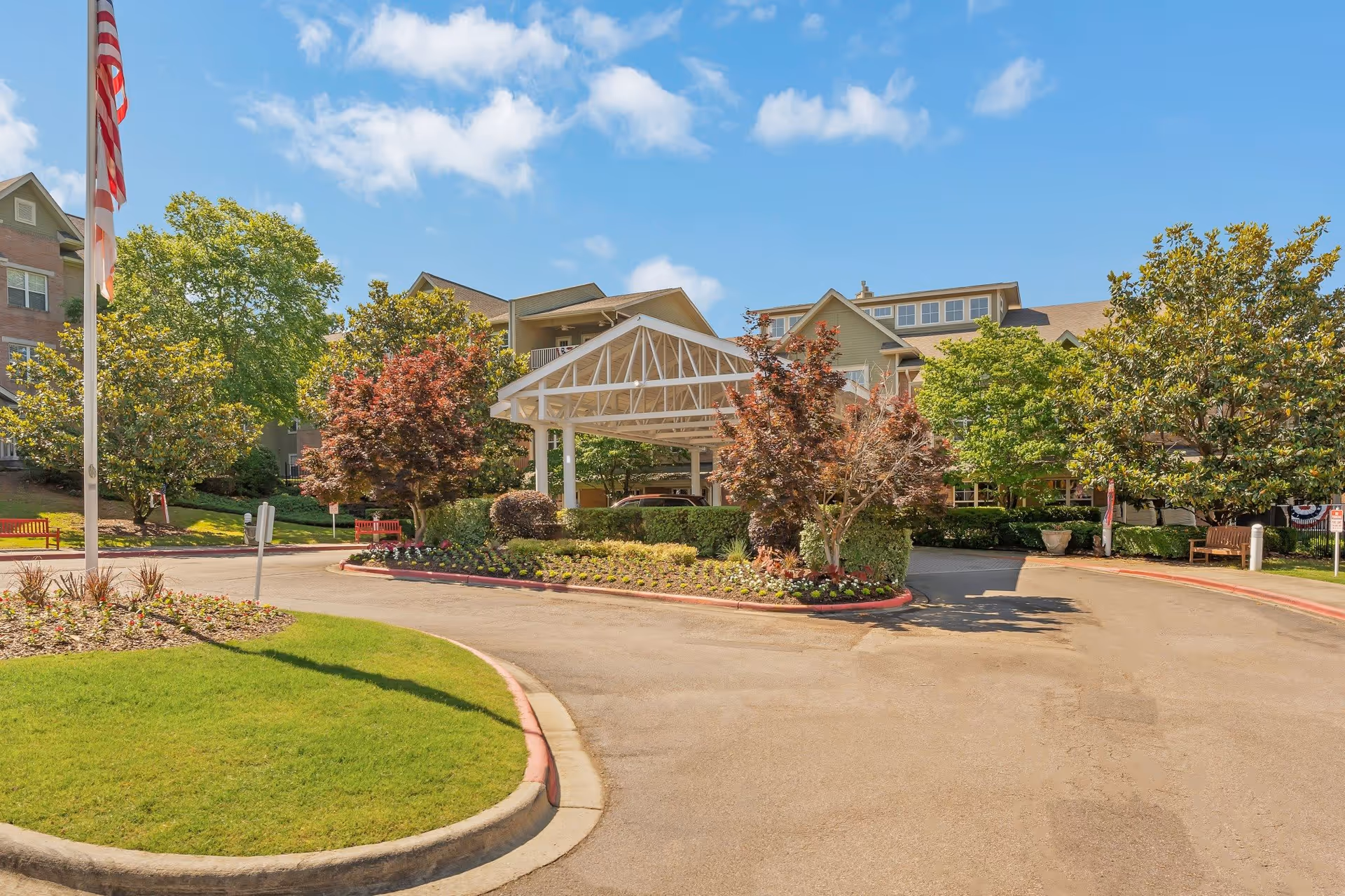 Entrance driveway to a senior living facility with a white covered drop-off area surrounded by landscaped greenery, trees, and flower beds under a blue sky with scattered clouds.