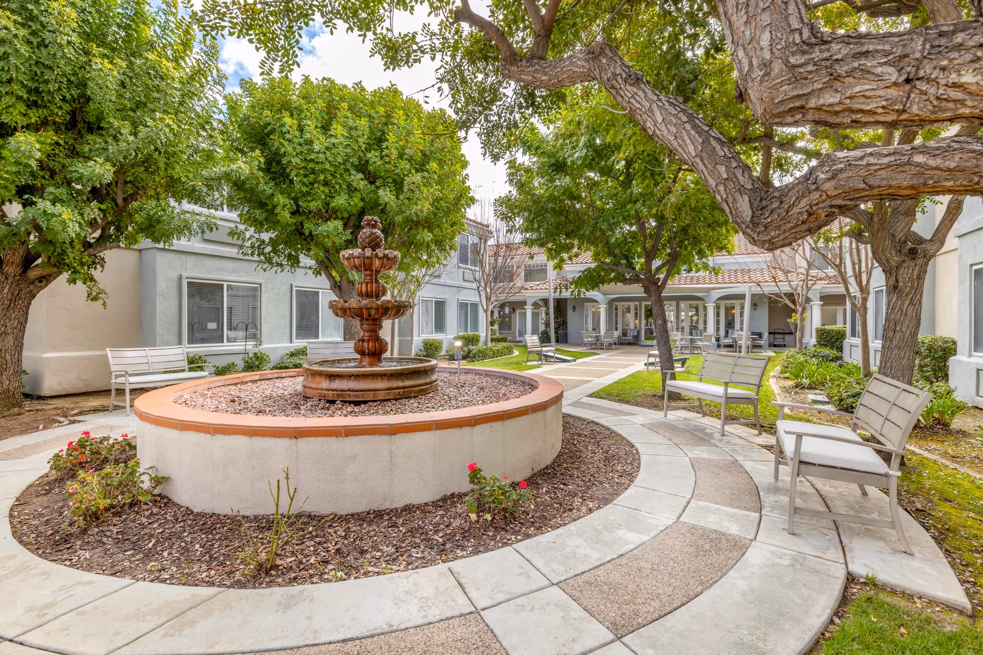 Outdoor courtyard area with a circular stone fountain in the center surrounded by trees, benches, and a paved walkway. The courtyard is enclosed by a light-colored building with windows and a covered patio area with chairs and tables.