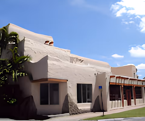 Exterior view of a white stucco building with a flat roof and rounded edges under a blue sky with some clouds. There is a small patch of grass and a sidewalk in front of the building.