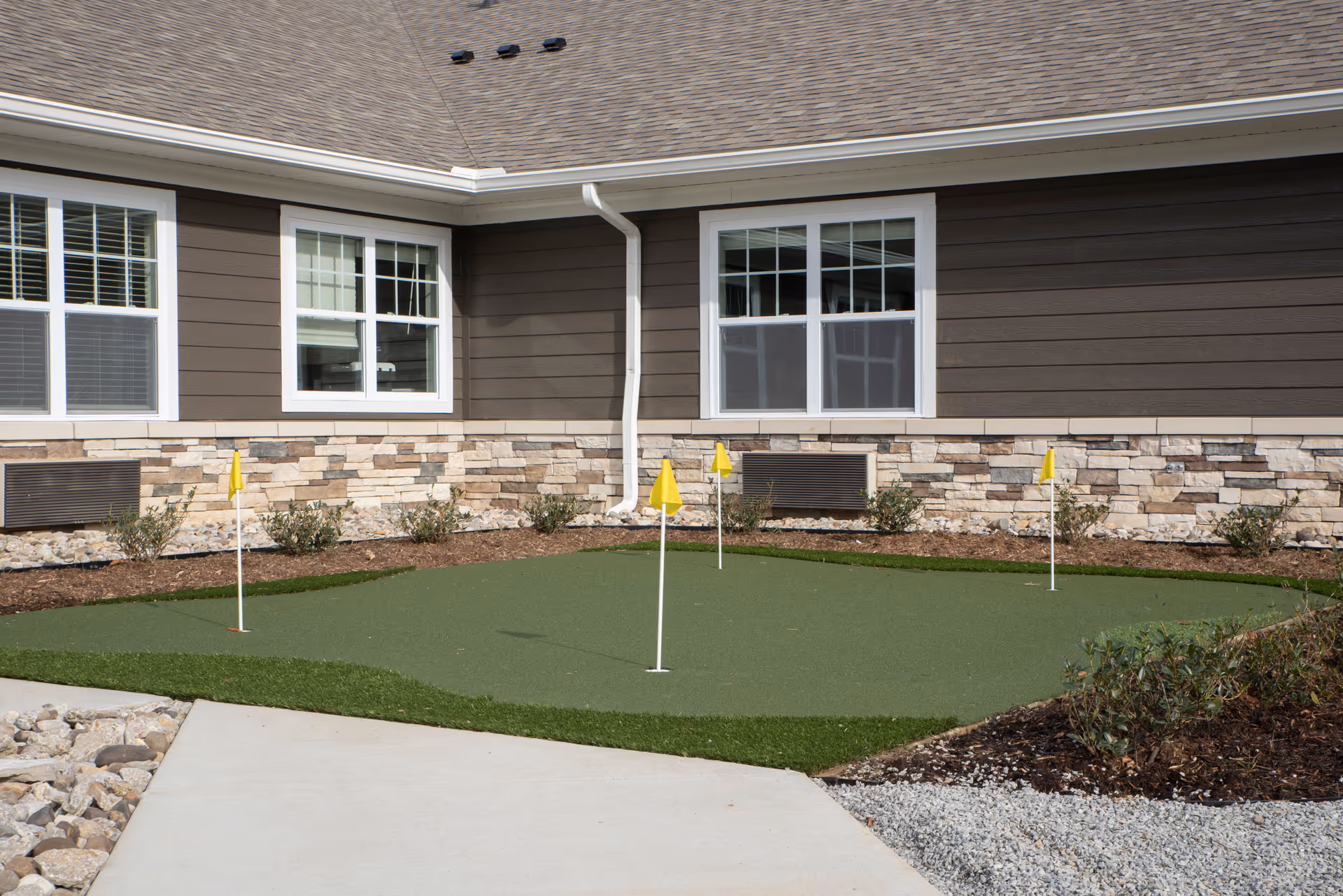 Outdoor putting green with four yellow flags in front of a building with brown siding and stone accents, surrounded by landscaping and a concrete walkway.