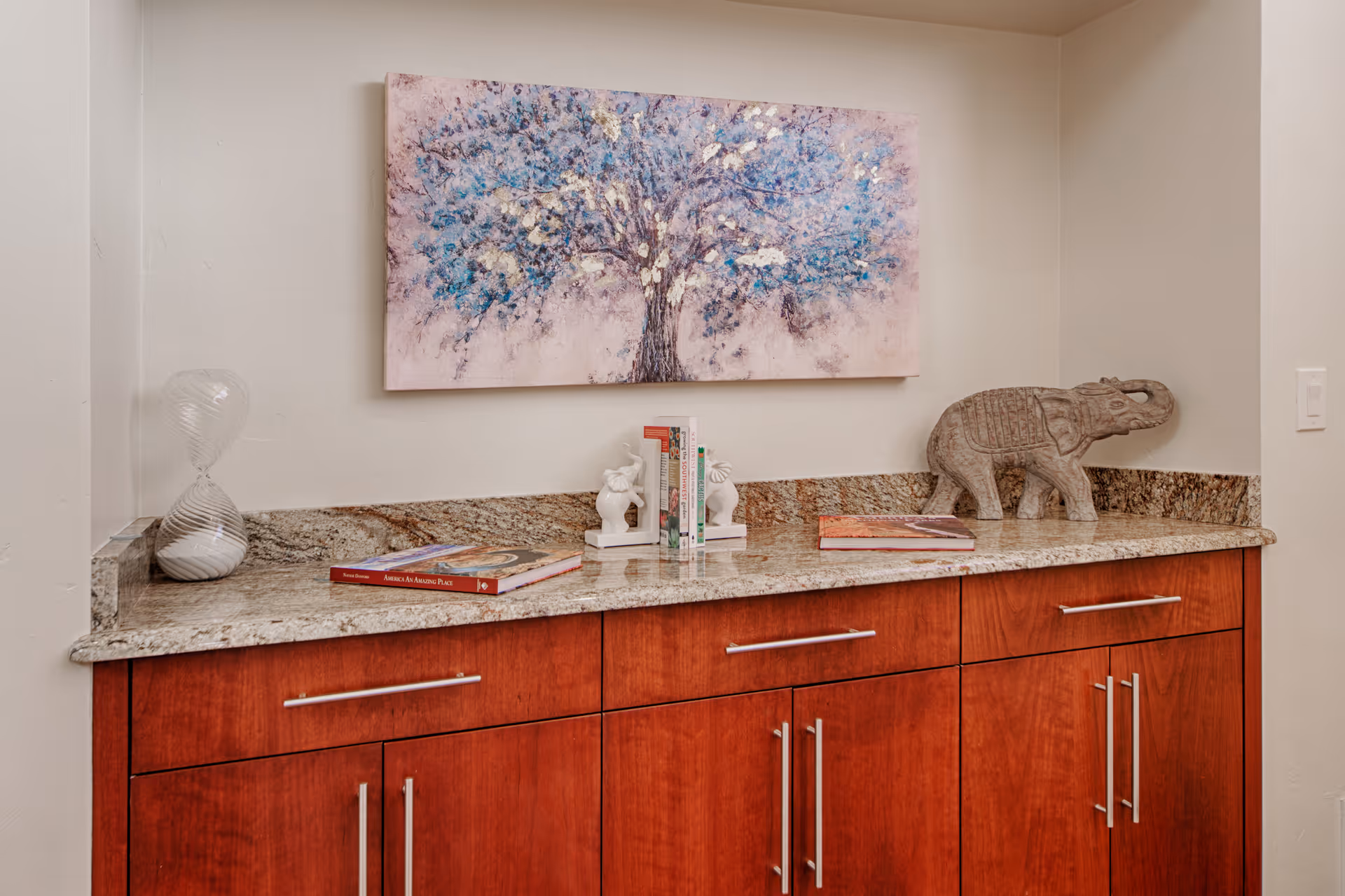 Granite-topped wooden cabinet with decorative books, sculptures and a tree painting on the wall above.