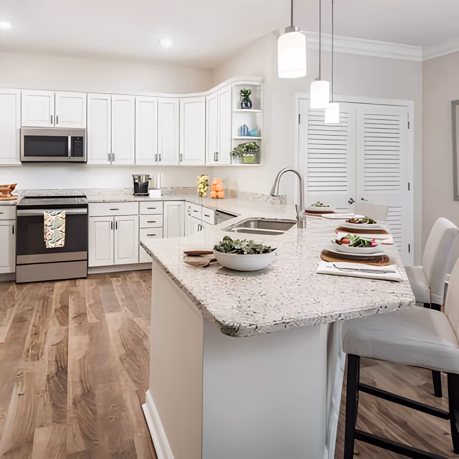 A modern kitchen with white cabinetry, a granite countertop island with a sink, and three pendant lights hanging above. The kitchen features stainless steel appliances including a microwave and stove. There are four place settings with plates of salad on the island, and wooden flooring throughout.