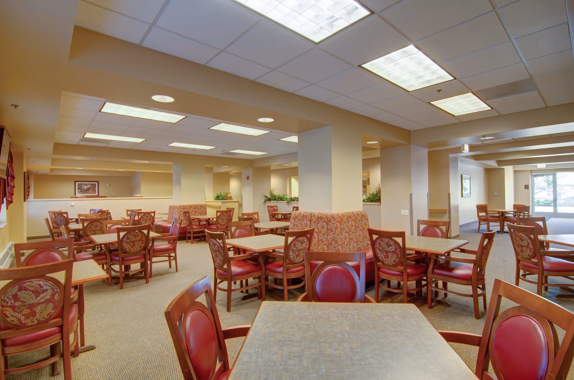 A spacious dining room with multiple wooden tables and chairs featuring red cushioned seats and floral-patterned backs. The room has beige walls, carpeted floors, and ceiling lights. There are some decorative plants and framed artwork on the walls, with large windows letting in natural light in the background.
