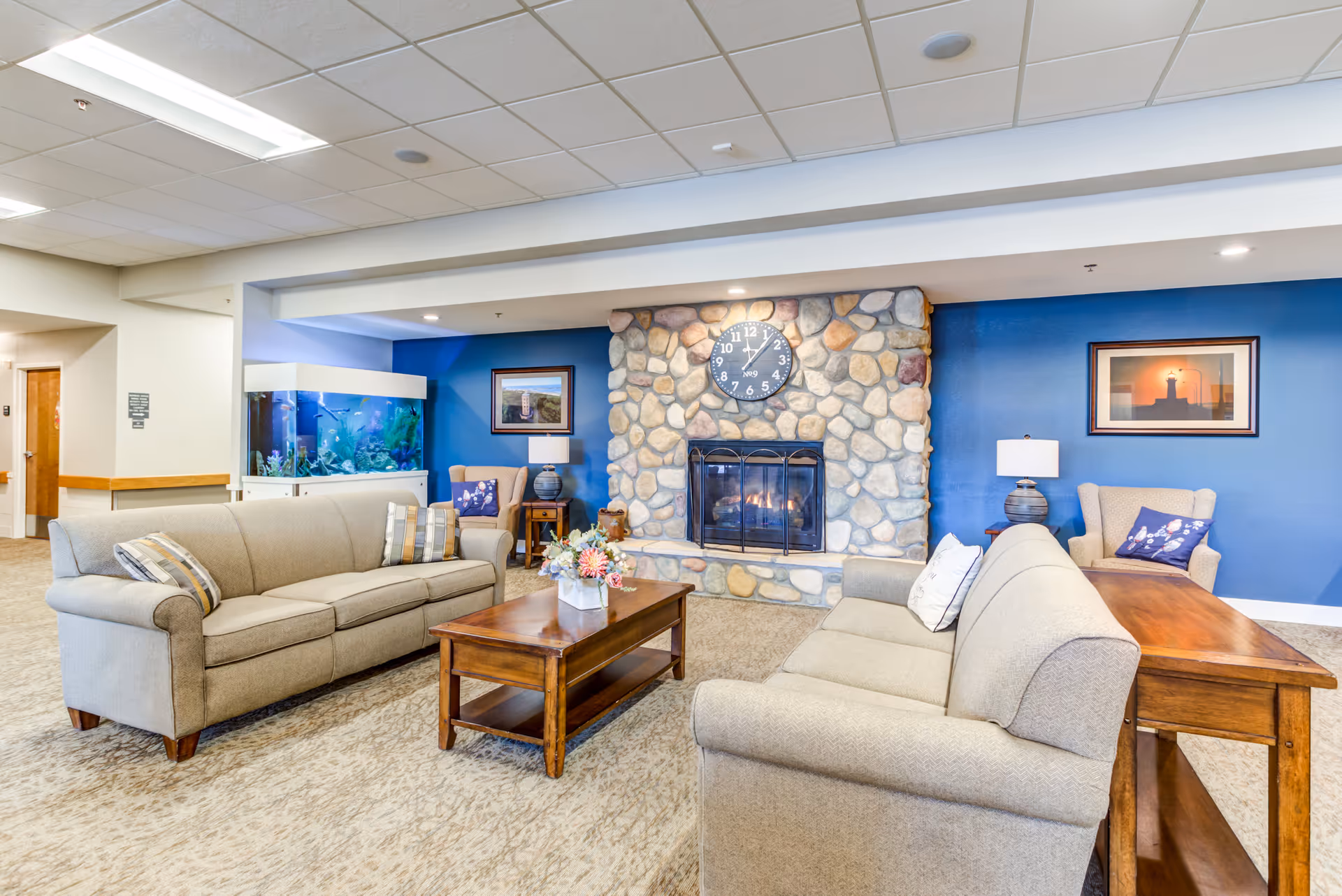 A cozy living room area in Keystone Bluffs Assisted Living featuring two beige sofas facing each other with a wooden coffee table in between. Behind one sofa is a wooden console table. The room has a stone fireplace with a clock above it on a blue accent wall. There are two armchairs with side tables and lamps, framed pictures on the walls, and a large fish tank in the corner.