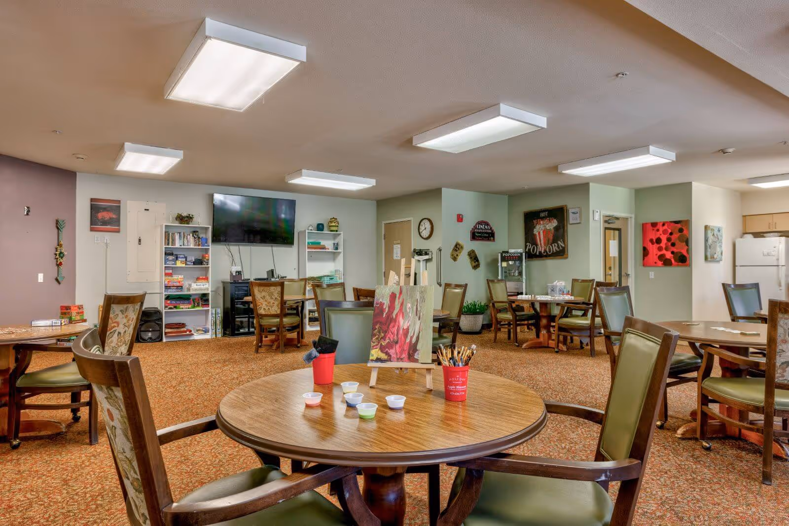 Spacious senior community dining/activity room with round tables and chairs, art supplies on a table, shelving and a wall-mounted TV.