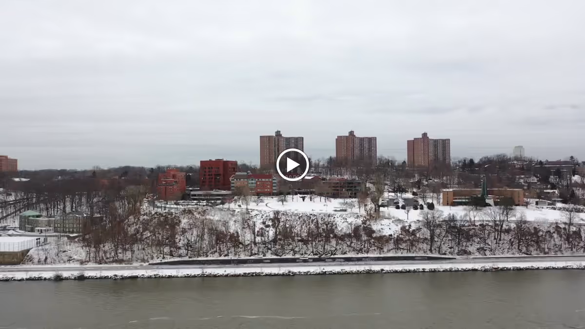 A snowy landscape showing a large facility complex with multiple brick buildings and high-rise towers situated on a hill overlooking a river under an overcast sky.