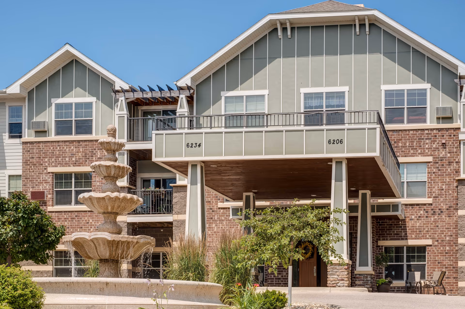 Exterior view of a multi-story senior living facility with a covered entrance supported by columns. The building features a combination of brick and light green siding with white trim. There is a decorative three-tiered stone fountain in the foreground surrounded by greenery and landscaping. The sky is clear and blue.