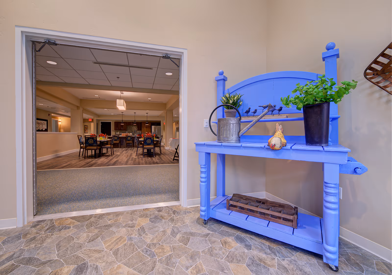 View into a communal dining area through an open doorway with a bright blue decorative bench holding plants and a watering can in the foreground.
