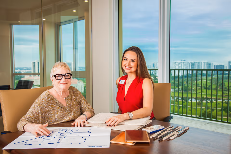 An elderly woman and a younger woman sitting at a table in a bright room with large windows showing a cityscape and greenery outside. The elderly woman is looking at floor plans spread out on the table, while the younger woman, wearing a red sleeveless top and a name tag, smiles at the camera. Various samples and materials are also on the table.