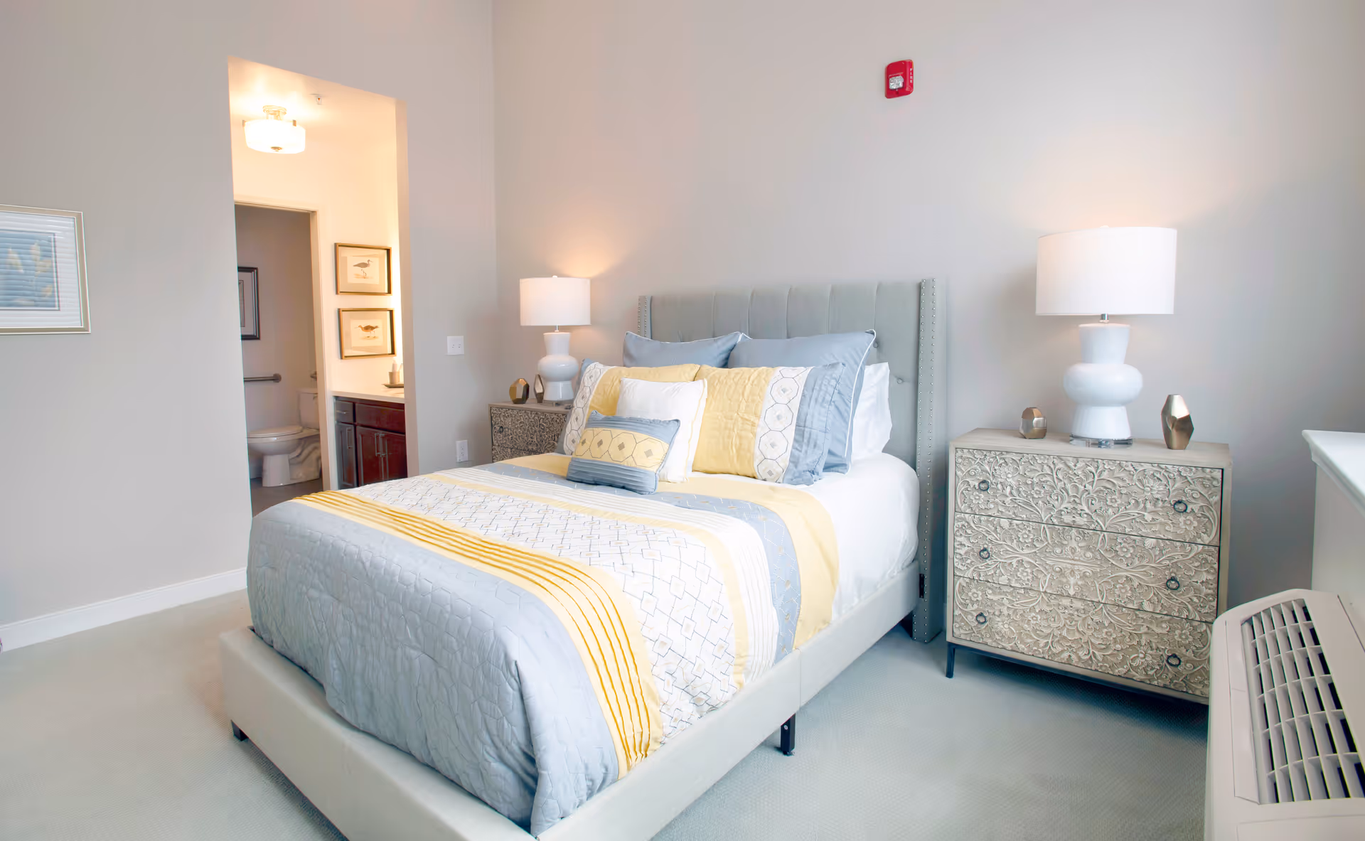 Well-lit bedroom with a bed dressed in gray and yellow bedding, two nightstands with lamps, and an open doorway to a bathroom.
