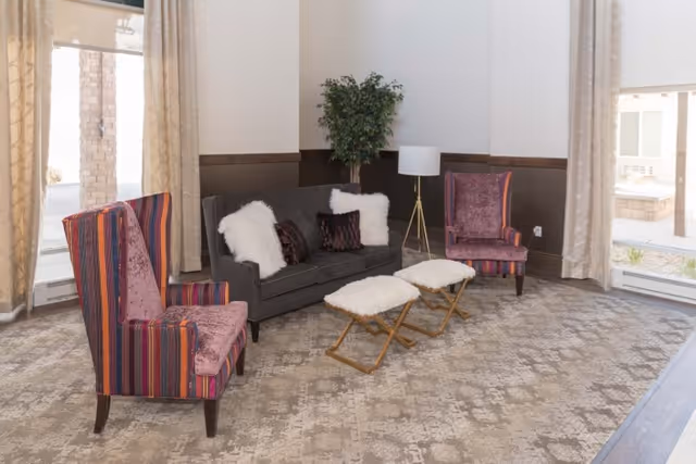 A cozy living room area with a dark gray sofa adorned with white and dark purple pillows, two colorful striped armchairs, two small cushioned stools with wooden legs, a floor lamp, and a potted plant in the corner. Large windows with beige curtains allow natural light to fill the room.
