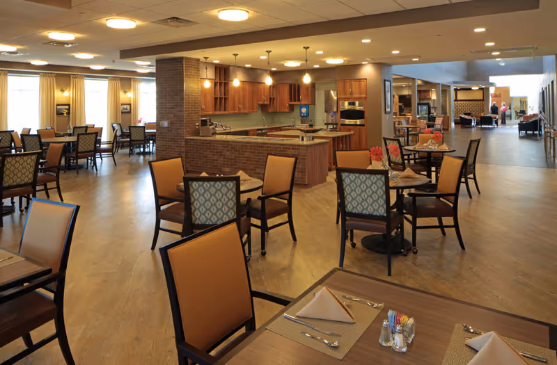 A spacious dining area in a senior living facility with multiple tables and chairs arranged neatly. The room features a kitchen area with wooden cabinets and modern appliances. The floor is wooden, and the space is well-lit with ceiling lights and natural light from windows with curtains.