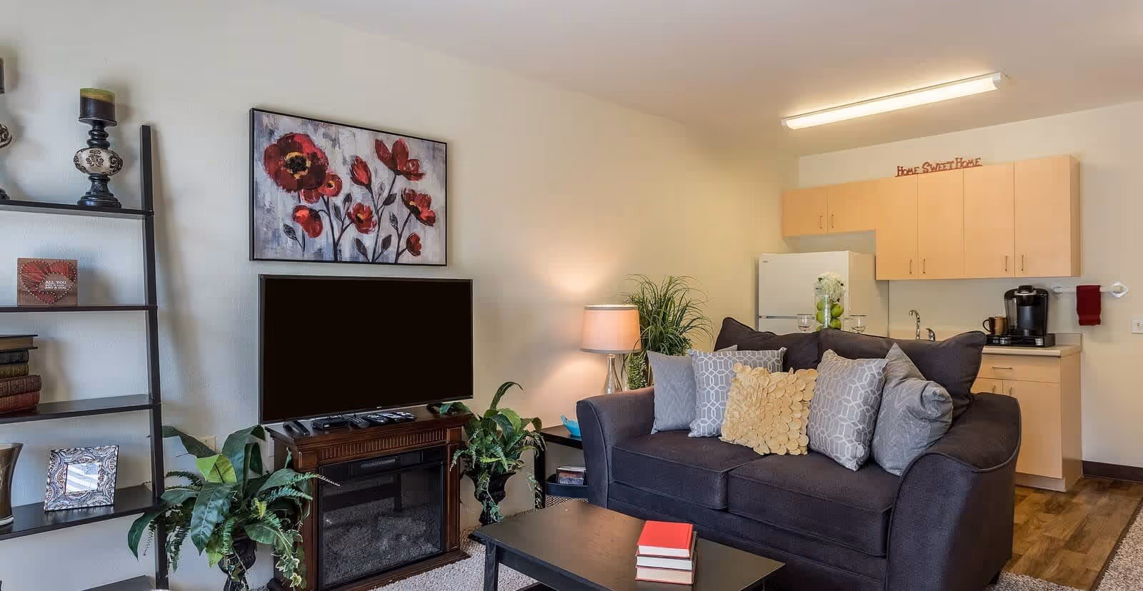 A cozy living room area with a dark gray sofa adorned with multiple decorative pillows, a black coffee table with two red books, a TV on a wooden stand with a built-in electric fireplace, and a tall black shelving unit with decorative items and plants. In the background, there is a small kitchen area with light wood cabinets, a white refrigerator, a coffee maker, and a sign above the cabinets that reads 'Home Sweet Home'.