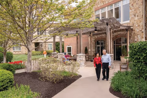 Two elderly people walking and talking on a paved pathway in a landscaped garden area outside a senior living community building with brick walls and large windows. Other residents are sitting on benches and at tables under a wooden pergola. Trees, bushes, and flowers surround the pathway.