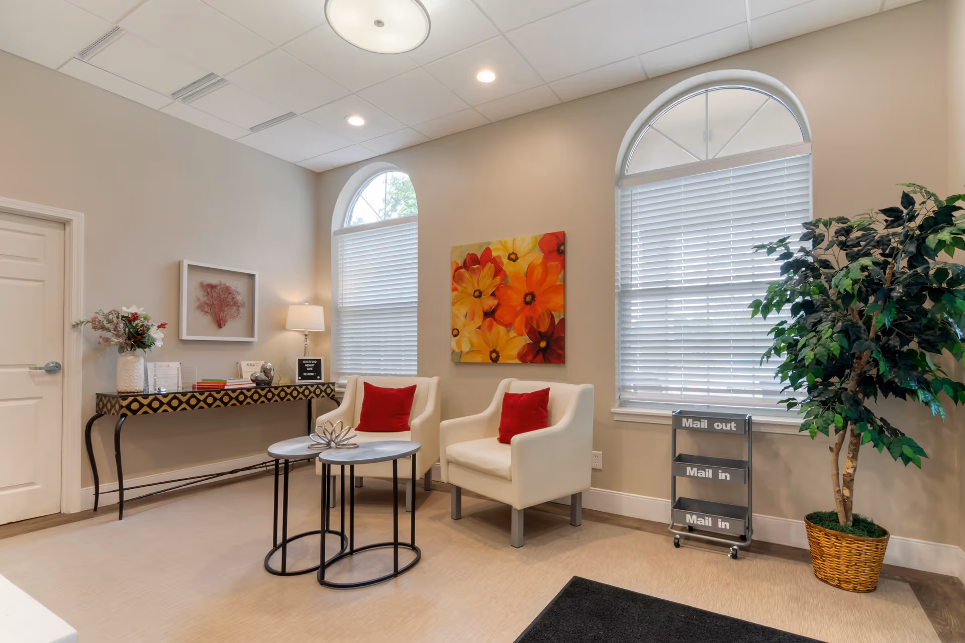 A cozy seating area in a memory care community with two white armchairs each having a red pillow, two nested round tables, a decorative console table with flowers and framed art above it, a large potted plant, and two arched windows with blinds. There is also a mail organizer labeled 'Mail out' and 'Mail in'.