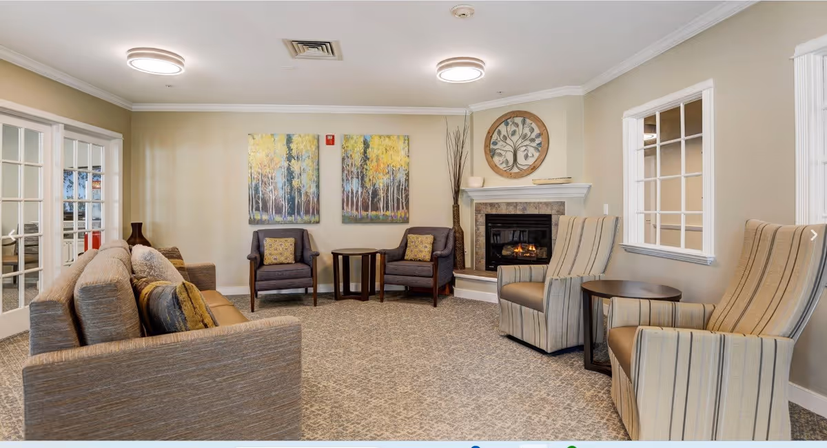 A cozy living room area with a beige carpet and neutral-colored walls. The room features a gray sofa with multiple cushions, two gray armchairs with patterned pillows, and two striped armchairs with small round tables beside them. A fireplace with a decorative round wall art above it is on one wall, and two paintings of trees hang on another wall. The room is well-lit with ceiling lights and has windows and glass-paneled doors.