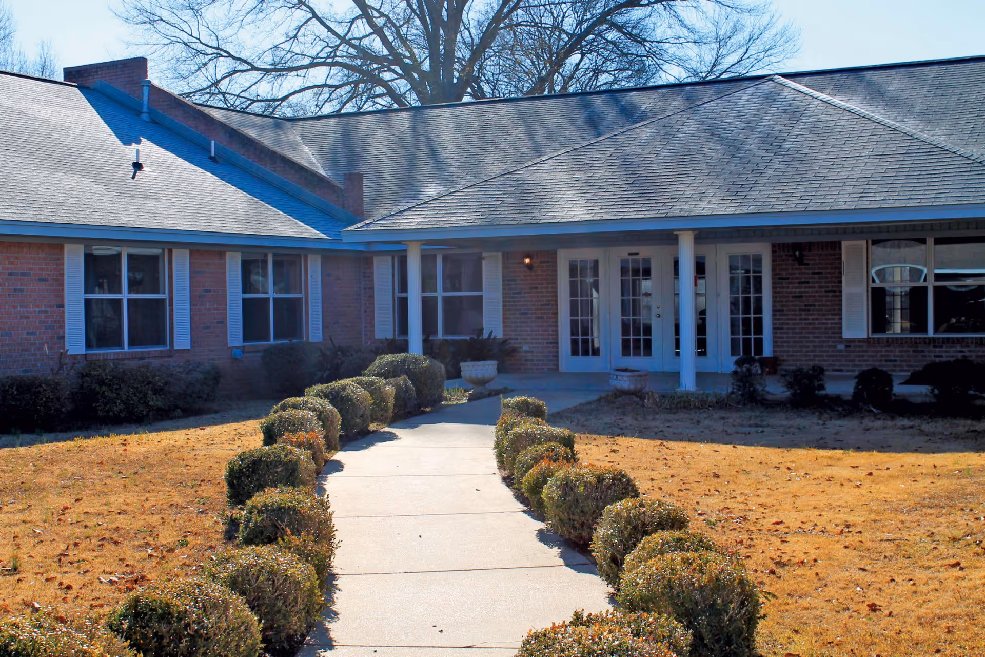 Front exterior view of a single-story brick building with a walkway lined by trimmed bushes leading to a covered entrance with white columns and glass doors. The lawn is dry and there are leafless trees in the background.