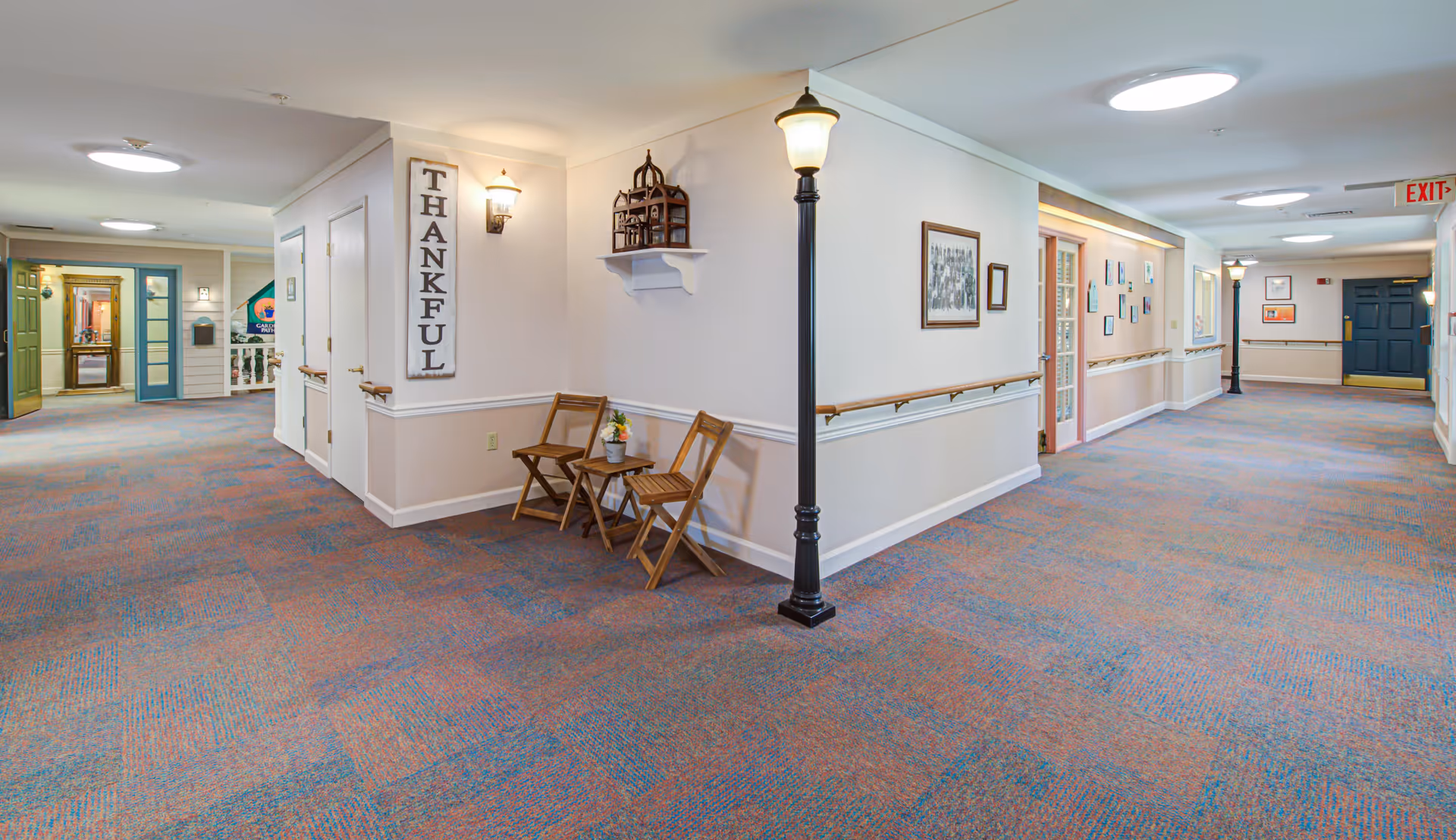 Wide carpeted interior hallway of a memory care facility with folding chairs, decorative lamp-post streetlights, wall handrails, and doors along the corridor.