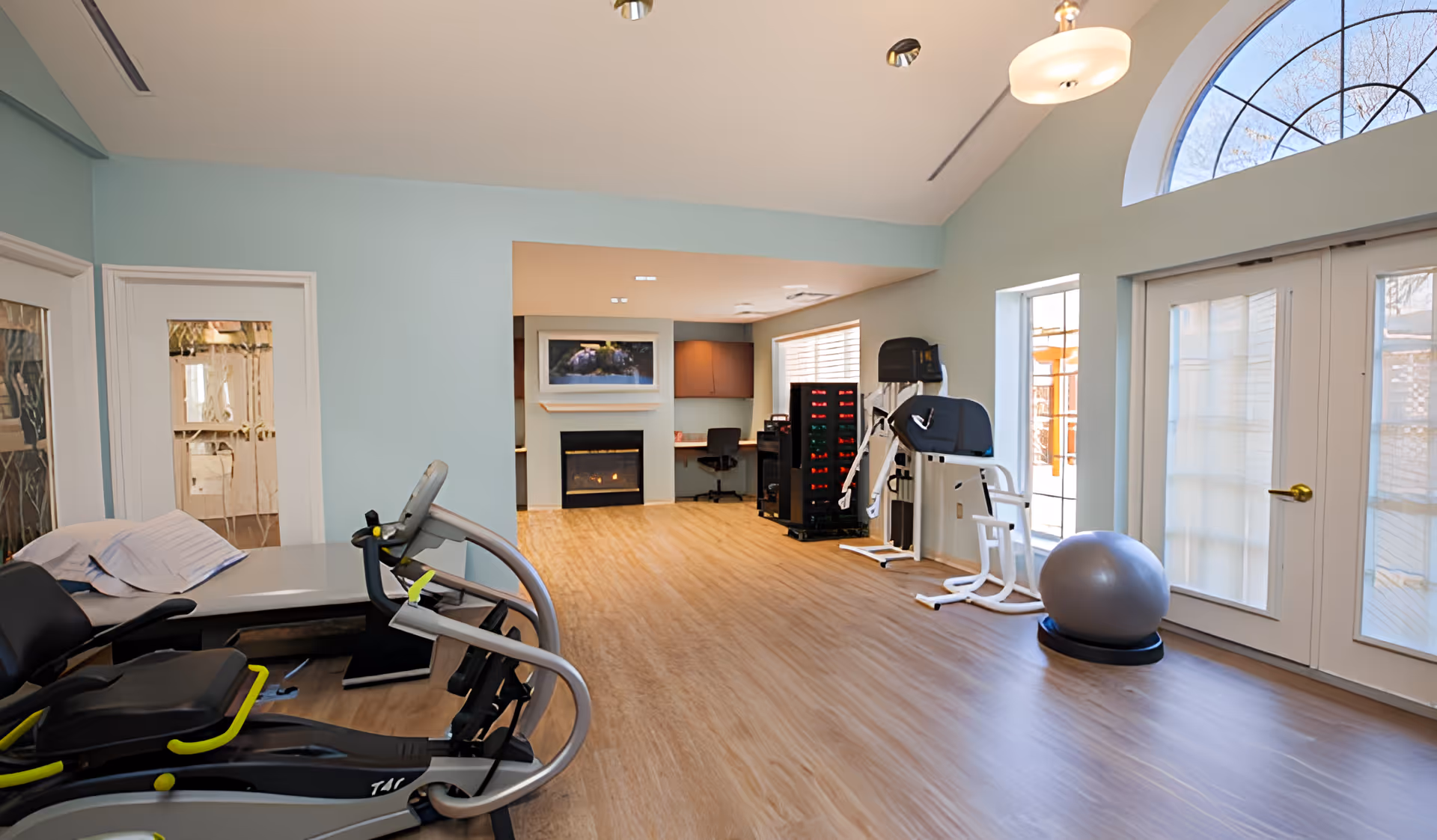 A bright exercise room with light blue walls and wood flooring. The room contains various exercise equipment including a recumbent bike, a multi-station gym machine, and an exercise ball. There is a fireplace with a TV mounted above it in the background, a desk with a chair, and large windows and glass doors letting in natural light.