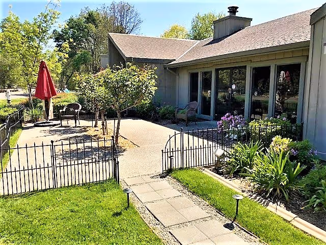 Outdoor patio and small fenced garden with a paved walkway, seating, and umbrella next to a single-story building.